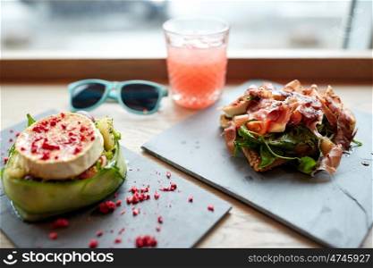 food, culinary, haute cuisine and cooking concept - goat cheese and prosciutto ham salads on stone plates with glass of juice and sunglasses on table at restaurant or cafe