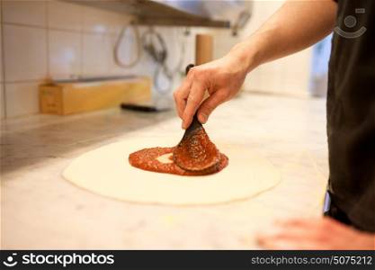 food, culinary, italian cuisine, people and cooking concept - cook with spoon applying tomato sauce to raw pizza dough at pizzeria. cook applying tomato sauce to pizza at pizzeria