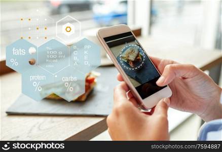 food, culinary, technology and people concept - close up of hands with panini sandwich on smartphone screen and nutritional value chart at restaurant. hands with phone and food nutritional value chart