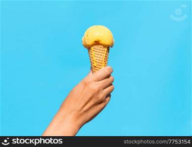 food, dessert and eating concept - close up of hand holding yellow orange ice cream in waffle cone over blue background. close up of hand holding waffle ice cream cone
