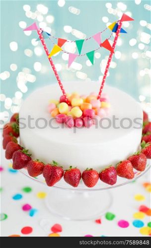 food, dessert and party concept - close up of birthday cake with candies, garland and strawberries on stand over lights on blue background. close up of birthday cake with garland on stand