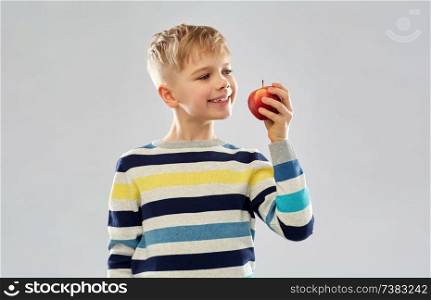 food, diet and children concept - smiling boy in striped pullover eating red apple over grey background. smiling boy in striped pullover eating red apple