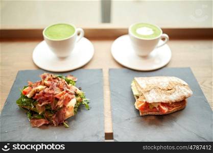 food, dinner, haute cuisine and eating concept - prosciutto ham salad with salmon panini sandwich on stone plates and cups of matcha green tea latte at restaurant