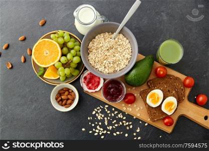 food, eating and breakfast concept - oatmeal cereals in bowl, fruits, almond nuts, glass of juice and jug with milk on slate stone table. oatmeal, fruits, toast bread, egg, jam and milk