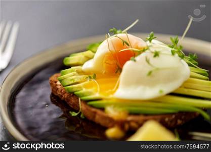 food, eating and breakfast concept - toast bread with sliced avocado, pouched egg and greens on ceramic plate. toast bread with sliced avocado and pouched egg