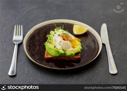 food, eating and breakfast concept - toast bread with sliced avocado, pouched egg and greens on ceramic plate with fork and knife. toast bread with sliced avocado and pouched egg
