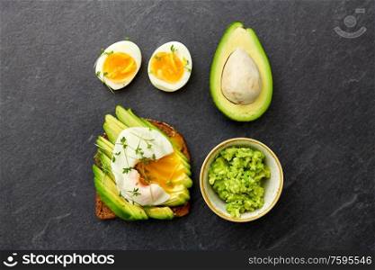 food, eating and breakfast concept - toast bread with sliced avocado, pouched egg and greens on slate stone table. toast bread with avocado, pouched egg and greens