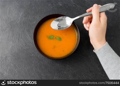 food, eating and new nordic cuisine concept - close up of hands with bowl of pumpkin cream soup and spoon on stone table. hands with bowl of pumpkin cream soup on table