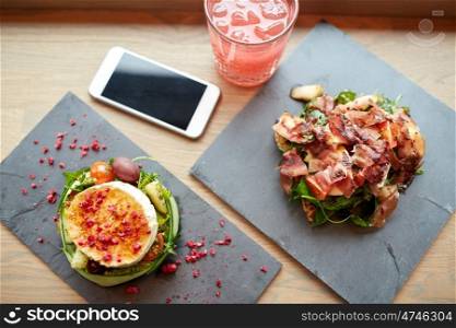 food, eating and technology concept - goat cheese and prosciutto ham salads on stone plates with smartphone and glass of drink at restaurant or cafe