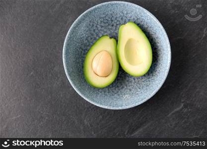food, eating and vegetable concept - close up of cut avocado with bone in ceramic bowl on slate stone background. close up of ripe avocado with bone in ceramic bowl