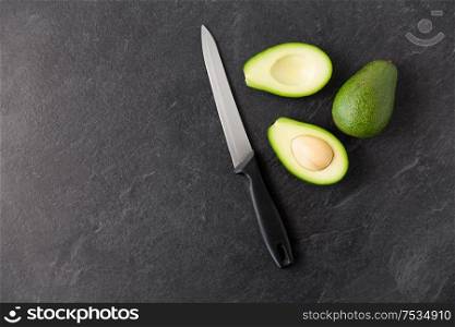food, eating and vegetable concept - close up of cut avocados with bone and kitchen knife on slate stone background. avocados and knife on slate stone background