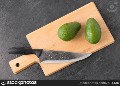 food, eating and vegetable concept - two avocados and kitchen knife on wooden cutting board l on slate stone background. two avocados and kitchen knife on cutting board