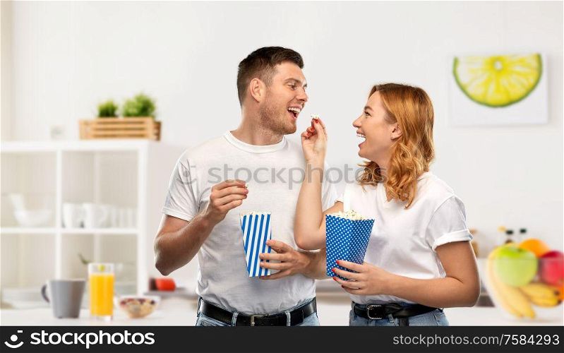 food, entertainment and people concept - portrait of happy couple in white t-shirts eating popcorn over home kitchen background. happy couple in white t-shirts eating popcorn