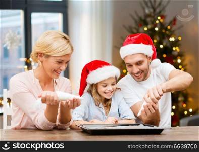food, family and holidays concept - happy mother, father and daughter family in santa hats decorating bakery by pastry bags with frosting over christmas tree lights background. family decorating bakery by frosting on christmas