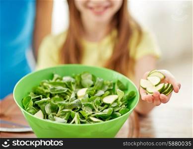 food, family, cooking and people concept - close up of happy girl making dinner and adding cucumber slices to green salad bowl in kitchen