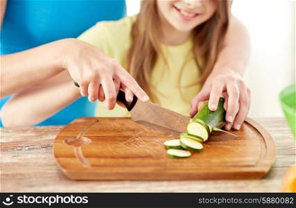 food, family, cooking and people concept - close up of happy girl and mother making dinner and chopping cucumber on cutting board with knife in kitchen