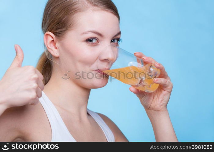 Food, health concept. Woman holding glass of orange flavored drink and drinking from it showing thumb up gesture. Studio shot on blue background. Woman drinking orange flavored drink or juice