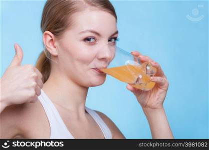 Food, health concept. Woman holding glass of orange flavored drink and drinking from it showing thumb up gesture. Studio shot on blue background. Woman drinking orange flavored drink or juice