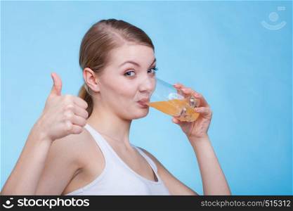 Food, health concept. Woman holding glass of orange flavored drink and drinking from it showing thumb up gesture. Studio shot on blue background. Woman drinking orange flavored drink or juice