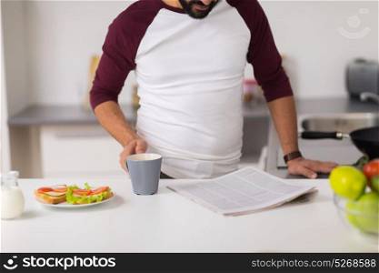 food, healthy eating and people concept - man having sandwiches with coffee for breakfast and reading newspaper at home kitchen. man reading newspaper and eating at home kitchen
