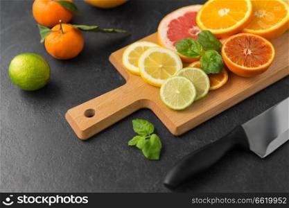 food, healthy eating and vegetarian concept - close up of citrus fruits, wooden cutting board and kitchen knife on slate table top. close up of fruits and knife on slate table top