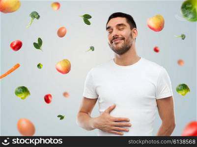 food, healthy eating, diet and people concept - happy full man touching his tummy over gray background with falling fruits