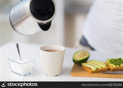 food, healthy eating, people and diet concept - kettle pouring hot water to tea cup and avocado sandwiches on table at home. kettle pouring hot water to tea cup at home