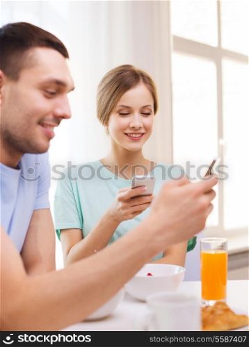 food, home, couple and technology concept - smiling couple with smartphones reading news and having breakfast at home