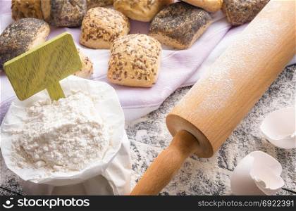 Food ingredients theme with a bag of flour with a blank wooden banner stick in it, a rolling pin and bread rolls baked from it, on a towel in the background.