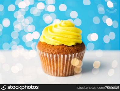 food, junk-food, culinary, baking and eating concept - close up of cupcake or muffin with yellow buttercream frosting over festive lights on blue background. close up of cupcake or muffin with yellow frosting