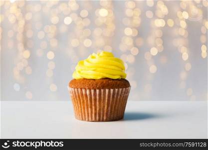 food, junk-food, culinary, baking and eating concept - close up of cupcake or muffin with yellow buttercream frosting over festive lights on background. close up of cupcake or muffin with yellow frosting