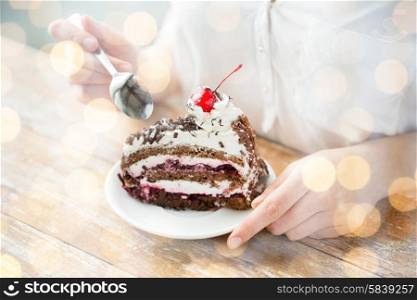 food, junk-food, culinary, baking and holidays concept - close up of woman eating chocolate cherry cake with spoon and sitting at wooden table over holidays lights background
