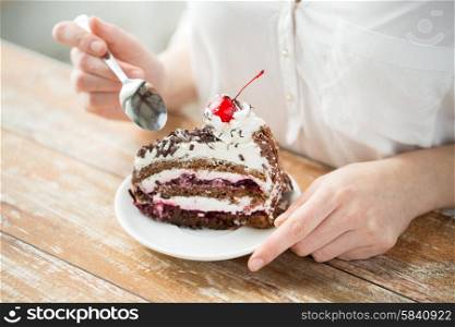 food, junk-food, culinary, baking and holidays concept - close up of woman eating chocolate cherry cake with spoon and sitting at wooden table