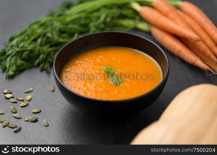 food, new nordic cuisine, culinary and cooking concept - close up of vegetable pumpkin cream soup with mint leaf in bowl, carrot and seeds on stone table. close up of pumpkin cream soup and vegetables