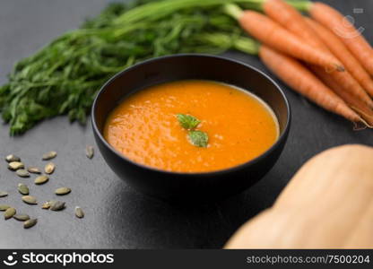 food, new nordic cuisine, culinary and cooking concept - close up of vegetable pumpkin cream soup with mint leaf in bowl, carrot and seeds on stone table. close up of pumpkin cream soup and vegetables