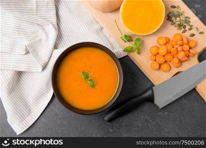 food, new nordic cuisine, culinary and cooking concept - close up of pumpkin cream soup in bowl, chopped vegetables and knife on stone table. close up of vegetable pumpkin cream soup in bowl
