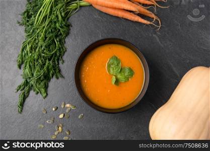 food, new nordic cuisine, culinary and cooking concept - close up of vegetable pumpkin cream soup with mint leaf in bowl, carrot and seeds on stone table. close up of pumpkin cream soup and vegetables