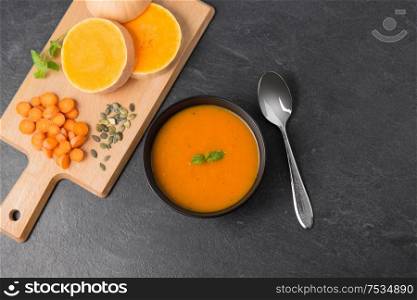 food, new nordic cuisine, culinary and cooking concept - close up of pumpkin cream soup in bowl, spoon and cutting board with vegetables on slate stone background. vegetable pumpkin cream soup in bowl with spoon