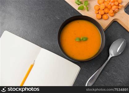 food, new nordic cuisine, culinary and cooking concept - close up of vegetable pumpkin cream soup in bowl, spoon and notebook with pencil on stone table. close up of pumpkin cream soup and notebook