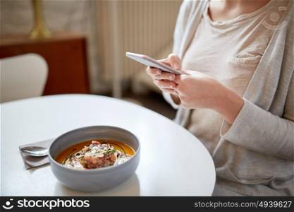 food, new nordic cuisine, technology, eating and people concept - woman sitting at cafe table with smartphone and bowl of vegetable pumpkin-ginger soup with goat cheese and tomato salad with yogurt. woman with smartphone and pumpkin soup at cafe