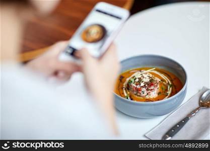 food, new nordic cuisine, technology, eating and people concept - woman with smartphone photographing bowl of vegetable pumpkin-ginger soup with goat cheese and tomato salad with yogurt at restaurant