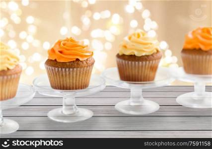 food, pastry and sweets concept - cupcakes with buttercream frosting on glass confectionery stand over festive lights background. cupcakes with frosting on confectionery stands