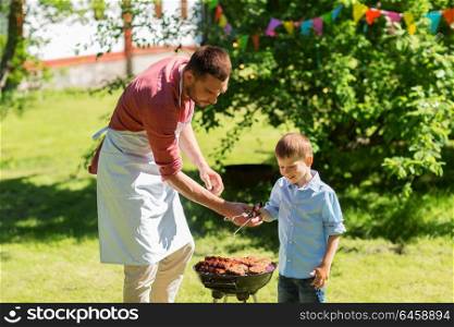 food, people and family time concept - father and son cooking meat on barbecue grill at bbq party in summer garden. father and son cooking meat on barbecue grill