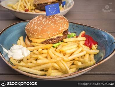 Food photography with a delicious big hamburger, french fries, mayonnaise, ketchup and a little European Union flag on top.