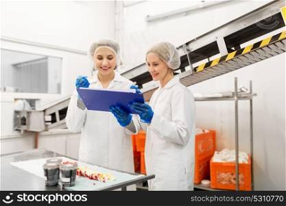 food production, industry and people concept - happy women technologists with clipboard tasting ice cream quality at factory. women technologists tasting ice cream at factory