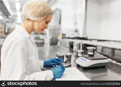 food production, industry and people concept - woman weighing ice cream on scale and filling papers at factory. woman working at ice cream factory