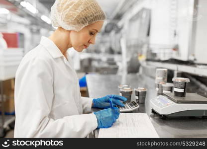 food production, industry and people concept - woman weighing ice cream on scale and filling papers at factory. woman working at ice cream factory