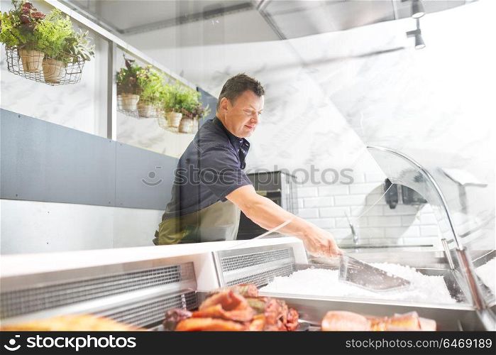 food sale, small business and people concept - male seller with scoop adding ice to fridge at fish shop. male seller adding ice to fridge at fish shop