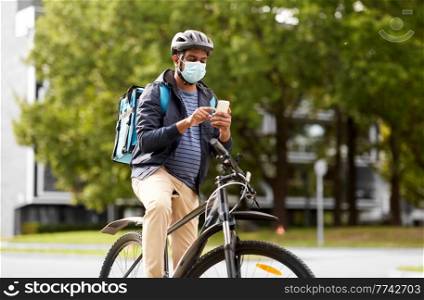 food shipping, health and people concept - indian delivery man in bike helmet and mask with thermal insulated bag and smatphone riding bicycle on city street. delivery man in mask with smatphone riding bicycle