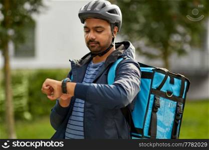 food shipping, profession and people concept - delivery man in bicycle helmet with thermal insulated bag looking at his wristwatch on city street. food delivery man looking at wristwatch in city